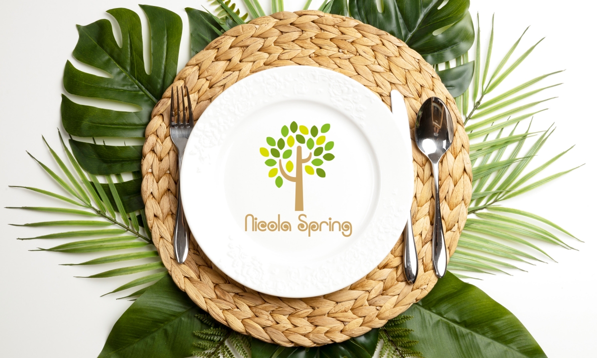 A white plate on a woven dried leaf placemat surrunded by leave and featuring a Nicola Spring logo on the plate